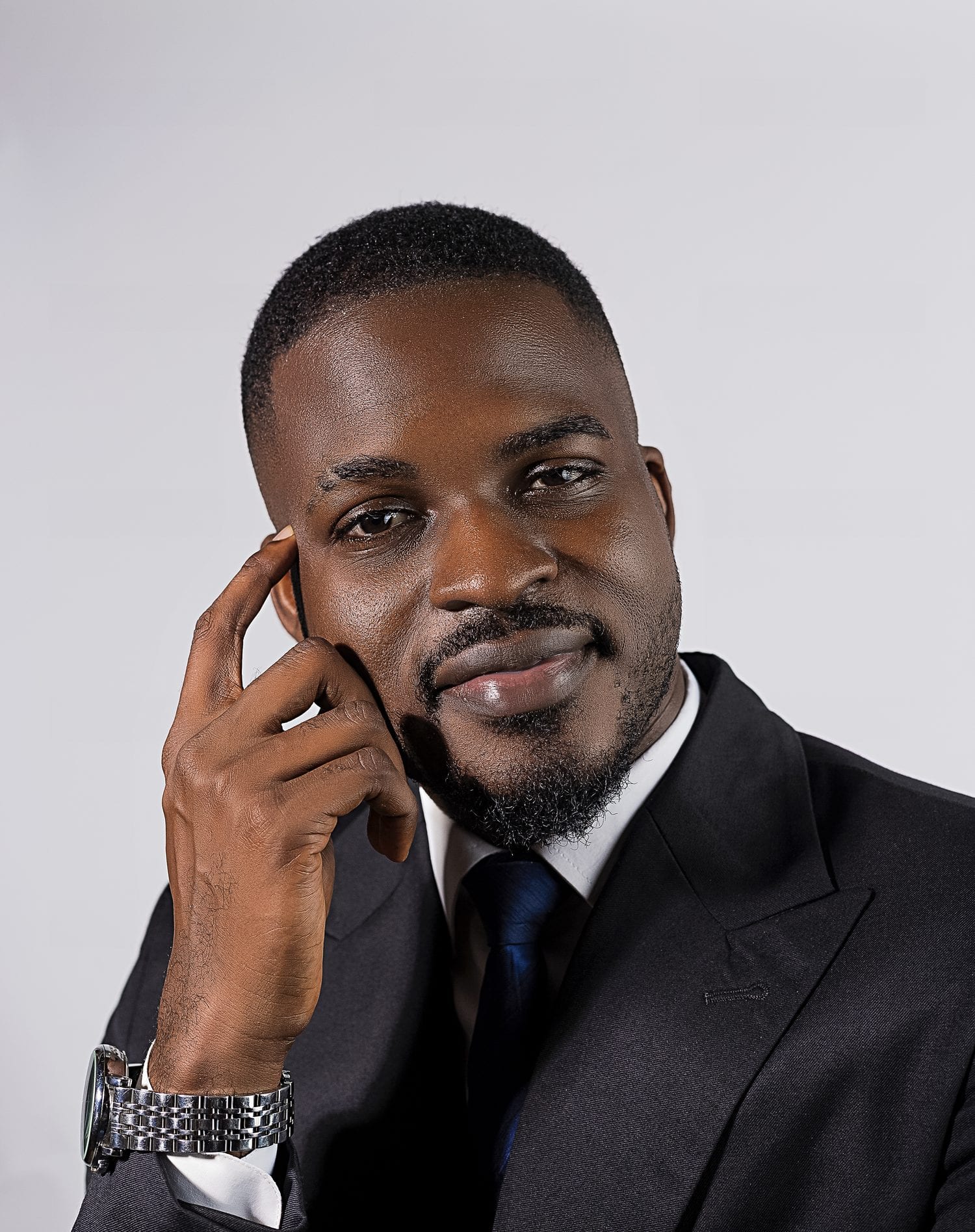 Tolu Afilaka in a dark suit, resting his hand against his face, looking to camera.